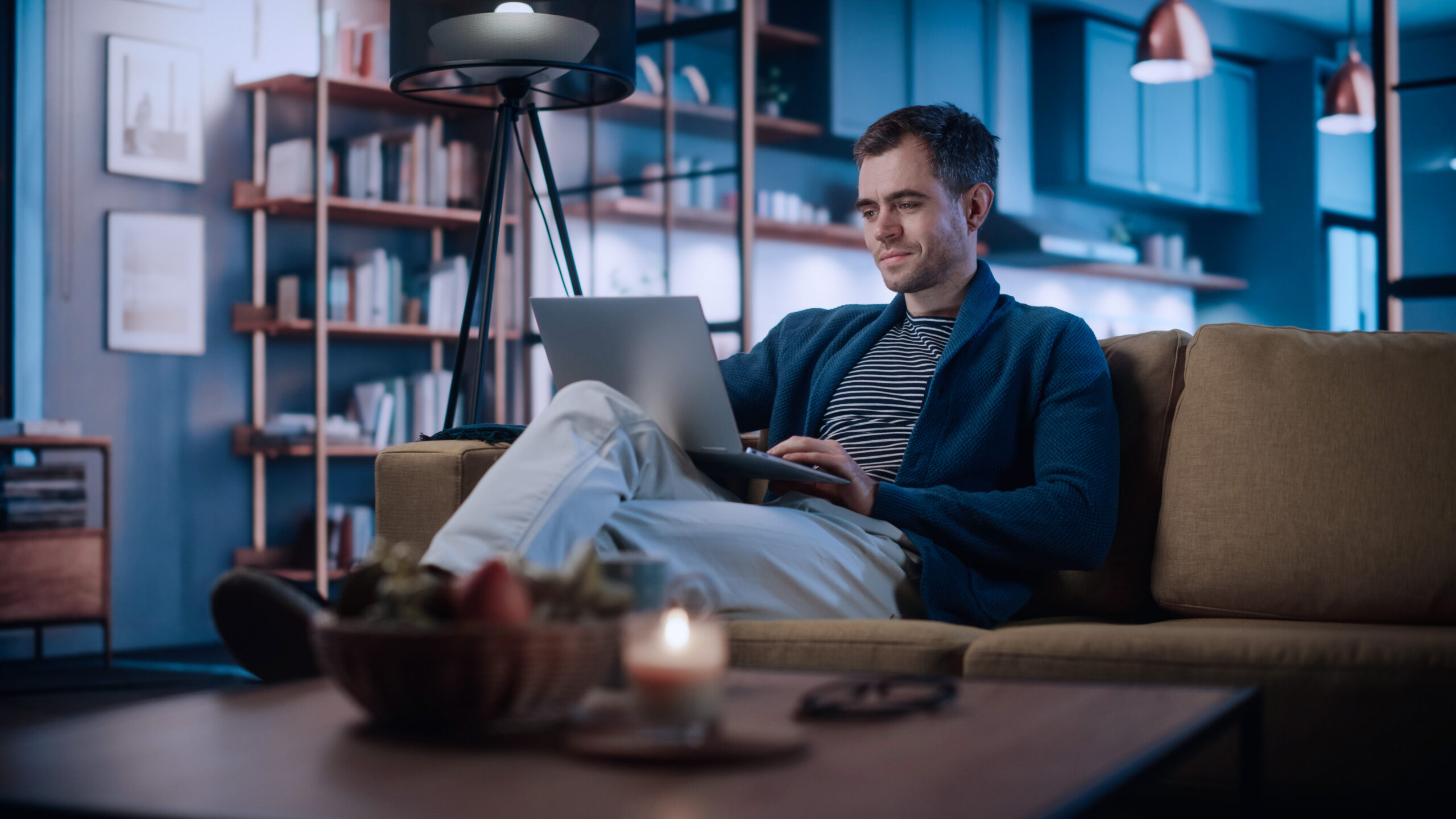 Photograph of a person sitting on a sofa using a laptop in a dimly lit living room with bookshelves and modern decor.