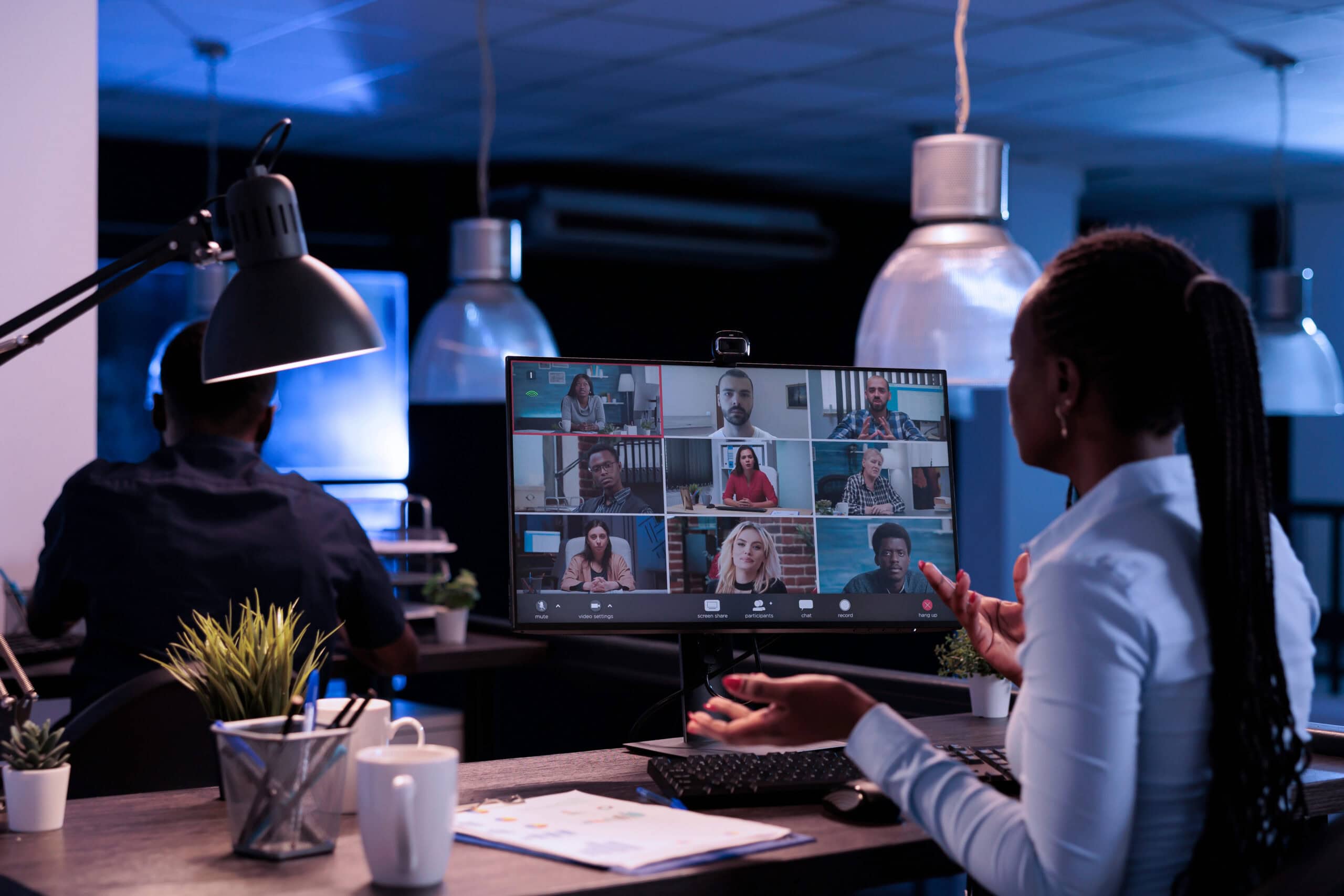 Photograph of a women in a modern office participating in a video conference call displayed on a computer monitor with nine participants in a grid layout.