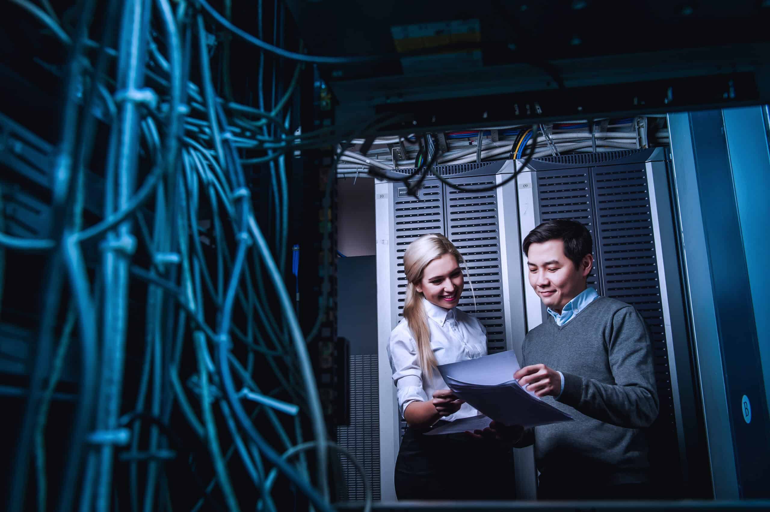 Photograph showing two IT professionals reviewing documents inside a server room surrounded by racks of network equipment and cables.